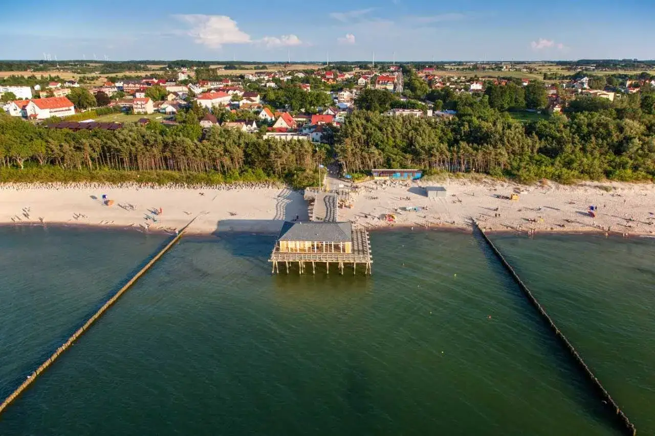 Urokliwe ustronie górskie nad morzem. Drewniany pomost z restauracją wchodzi w głąb turkusowej wody, a za nim rozciąga się piaszczysta plaża z wypoczywającymi ludźmi i sosnowy las.