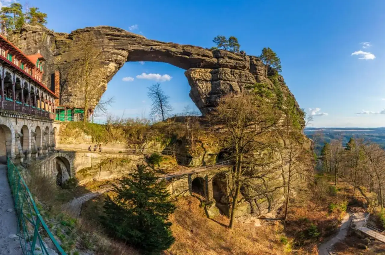 Imponujący skalny most w Czechach, z zabytkowym budynkiem po lewej i malowniczym krajobrazem w tle.