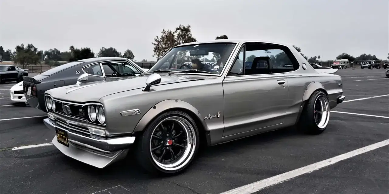 Srebrny Nissan Skyline z lat 70. z szerokimi błotnikami i czarnymi felgami. W tle widać inne klasyczne samochody, co pokazuje, jaki ruch jest w Japonii wśród fanów motoryzacji.