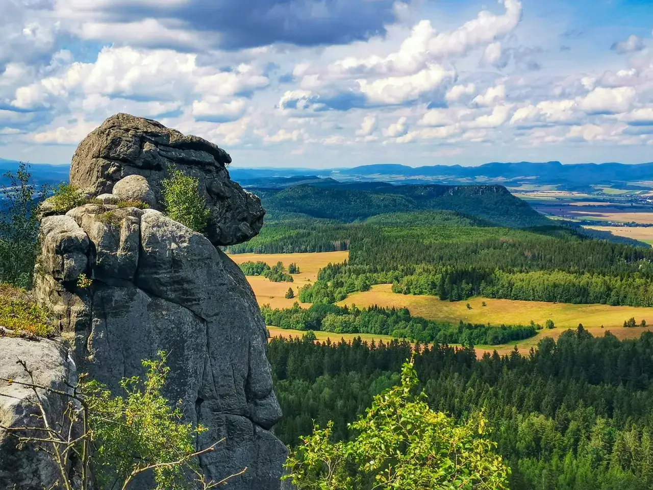 Skalny blok na pierwszym planie, z którego rozciąga się widok na zielone doliny i pagórki Gór Stołowych. Najwyższy szczyt w oddali.
