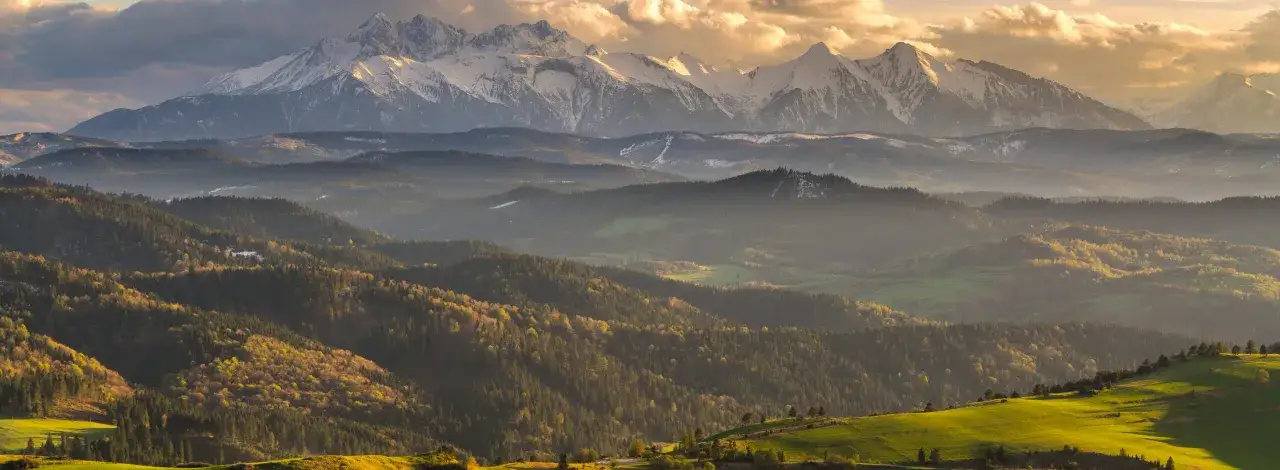 Złote światło oświetla ośnieżone szczyty górskie i pofałdowane, zalesione wzgórza. Idealne miejsce, by pomyśleć, gdzie w góry wybrać się na wędrówkę.