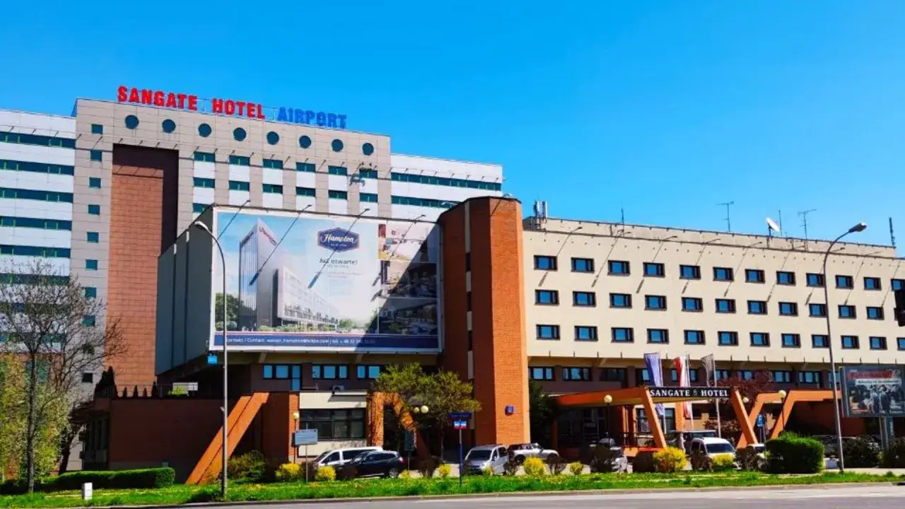 Hotel Sangate Airport, gotowy na Twój self check-in. Nowoczesny budynek z wieloma oknami i dużym banerem reklamowym.