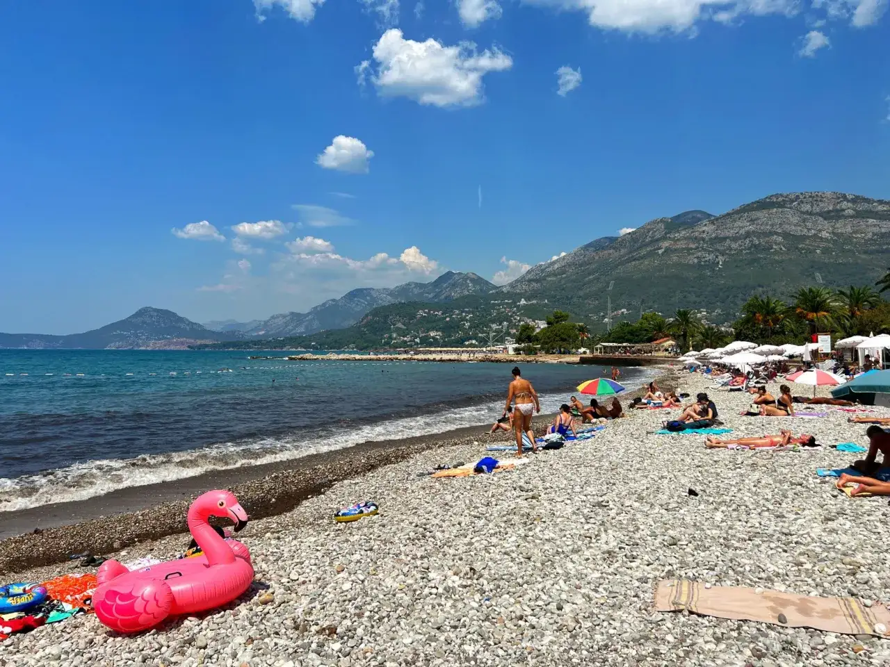 Słoneczna plaża w Czarnogórze z dziećmi. Różowy flaming unosi się na kamienistym brzegu, a w tle widać góry i turkusowe morze.