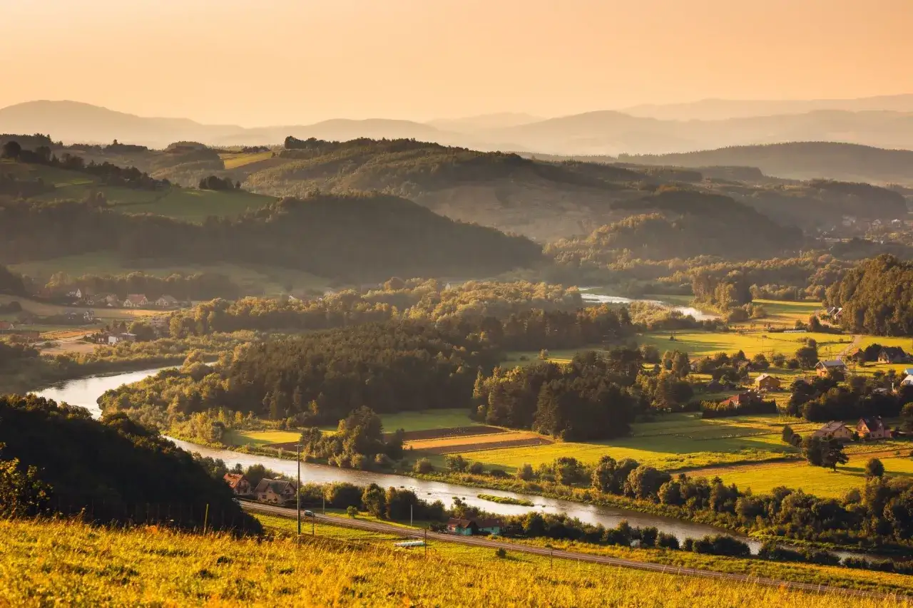 Widok na falujące wzgórza i dolinę rzeki o zachodzie słońca. W oddali widać kolejne pasma górskie. Krynica-Zdrój jakie to góry? To Beskid Sądecki.