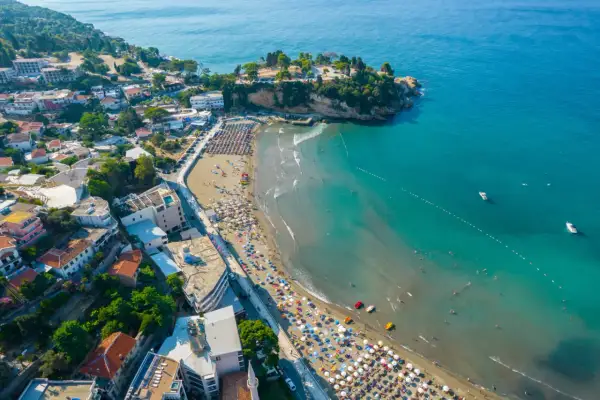 Widok na plażę w Czarnogórze z mnóstwem parasoli i kąpiącymi się ludźmi. W tle widać miasteczko i turkusowe morze. Idealne miejsce na wakacje z dziećmi.