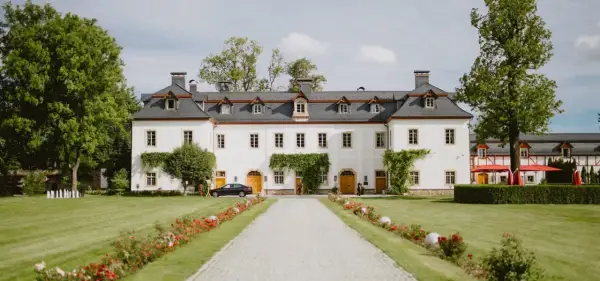 Biały pałac z szarym dachem, otoczony zielonym ogrodem z kwitnącymi kwiatami i drzewami.