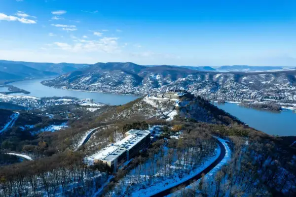 Zimowy krajobraz z górującym nad Dunajem **Wyszehrad**em. Na pierwszym planie hotel, w oddali zamek i ośnieżone wzgórza.