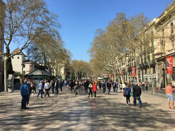 Tłum ludzi spaceruje po słynnej alei La Rambla w Barcelonie, otoczonej drzewami i budynkami.